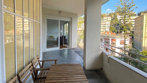 A balcony with a wooden table and benches, a glass door leading to an interior space, and a view of buildings and trees. - Penthouse near the beach and bike lane (Sanremo)