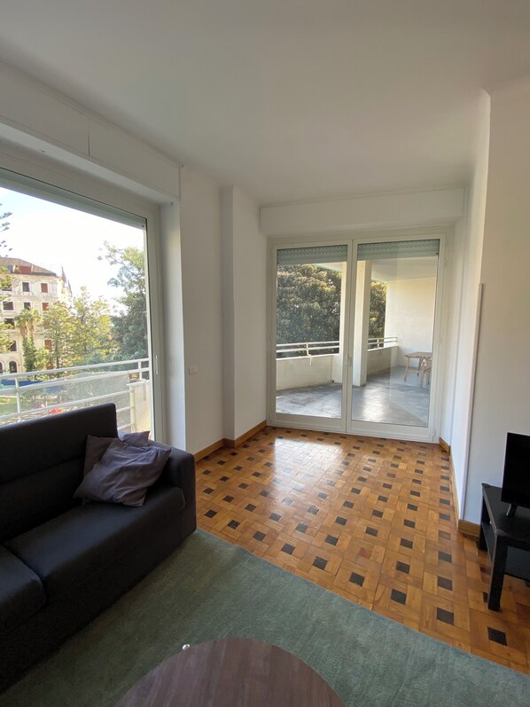 Amplio salón con un ventanal grande, piso de parquet de madera, un sofá oscuro y una mesita redonda.