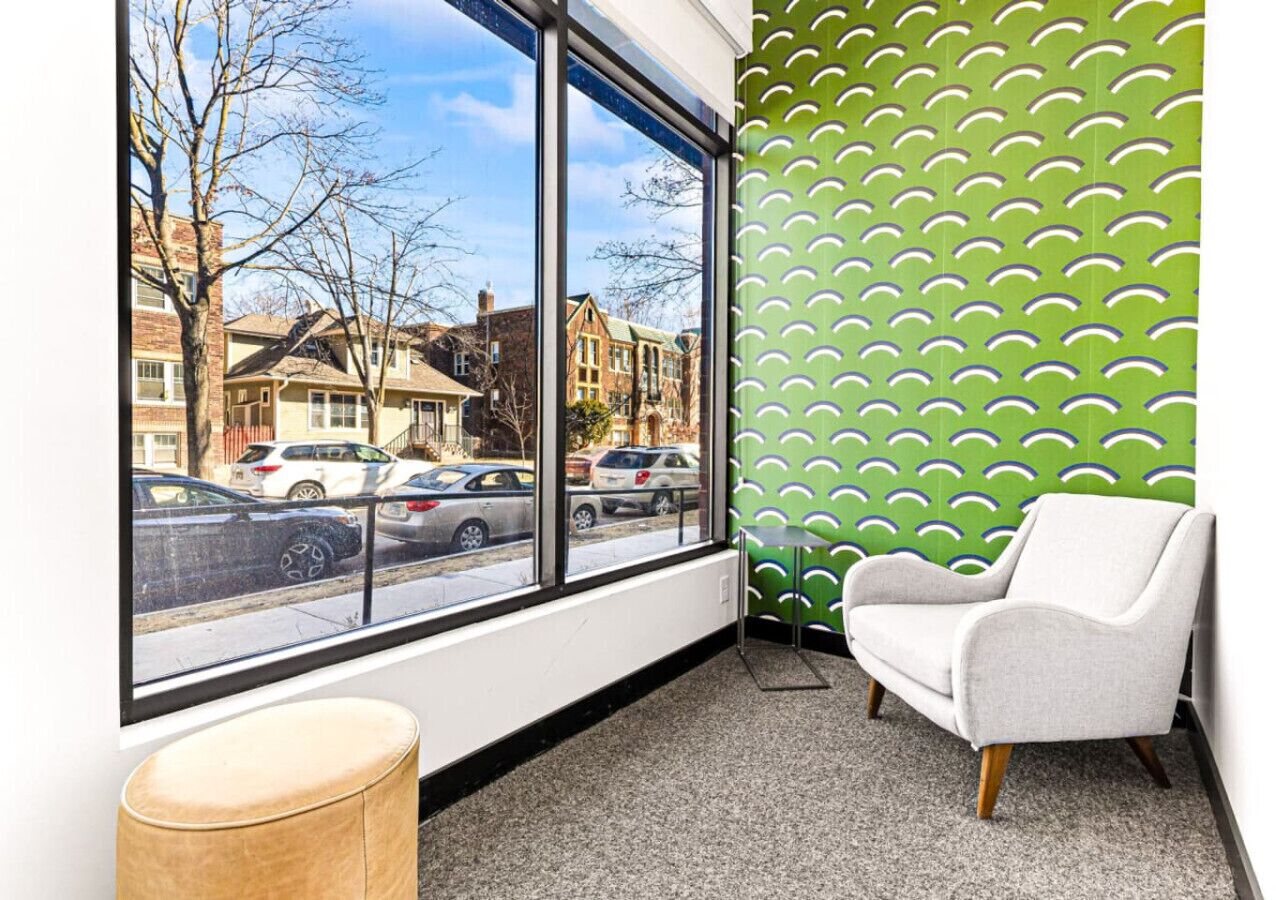 Une pièce avec une grande fenêtre, un mur orné d’un motif vert, un fauteuil blanc et un petit tabouret en bois.