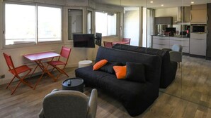 A modern living room with a black sofa, orange cushions, a small round table, and red folding chairs. A flat-screen TV is mounted on the wall, and a kitchen area is visible in the background. - Apartment in Grau-du-roi With Beach Access (Le Grau-Du-Roi)