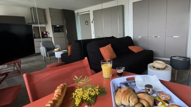 A modern living room with a black sofa, a red table with breakfast items, and a TV in the background. - Apartment in Grau-du-roi With Beach Access (Le Grau-Du-Roi)