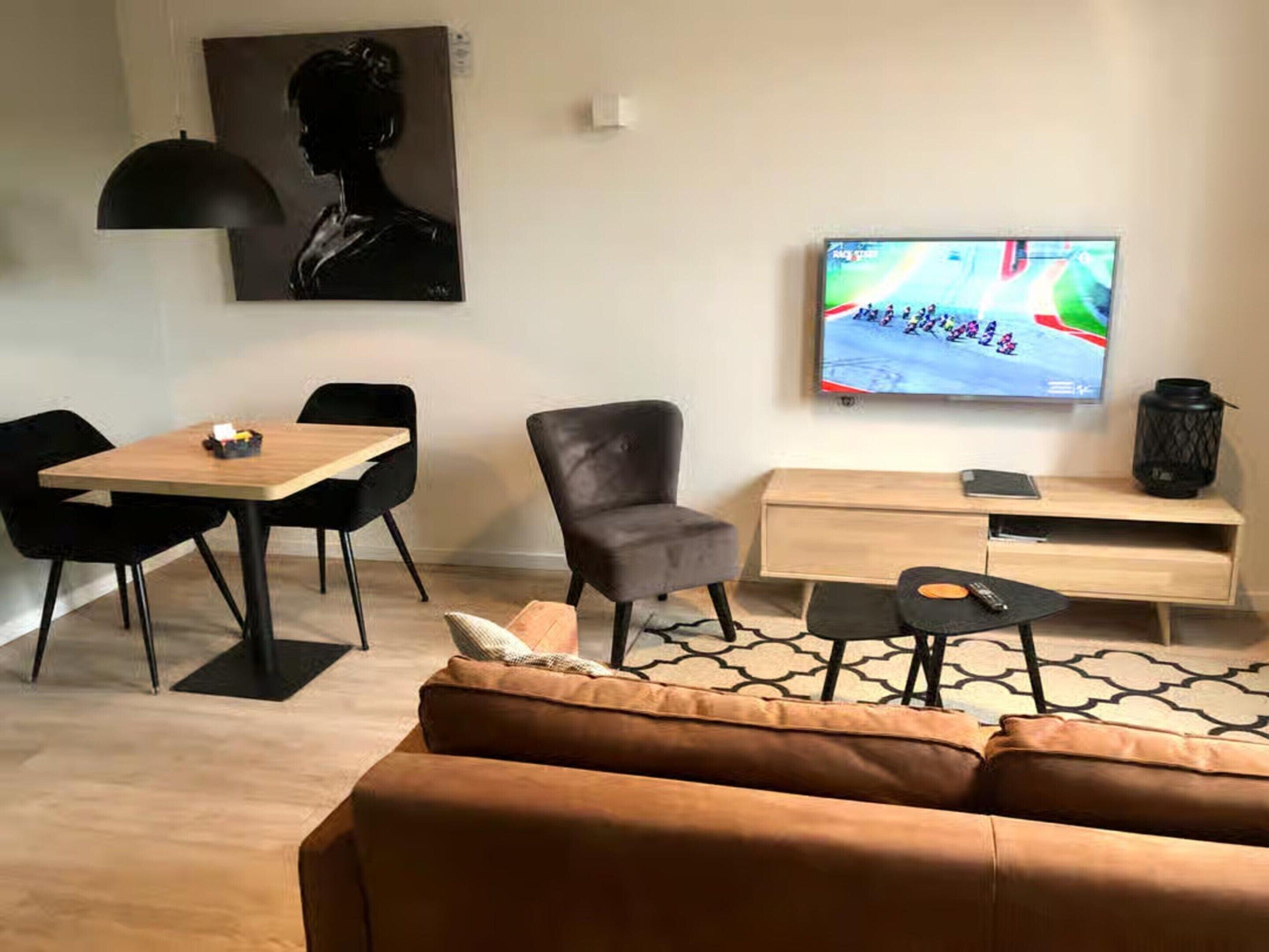Un salon moderne avec une table à manger en bois, des chaises noires, un canapé gris, un téléviseur fixé au mur et un tapis à motifs.