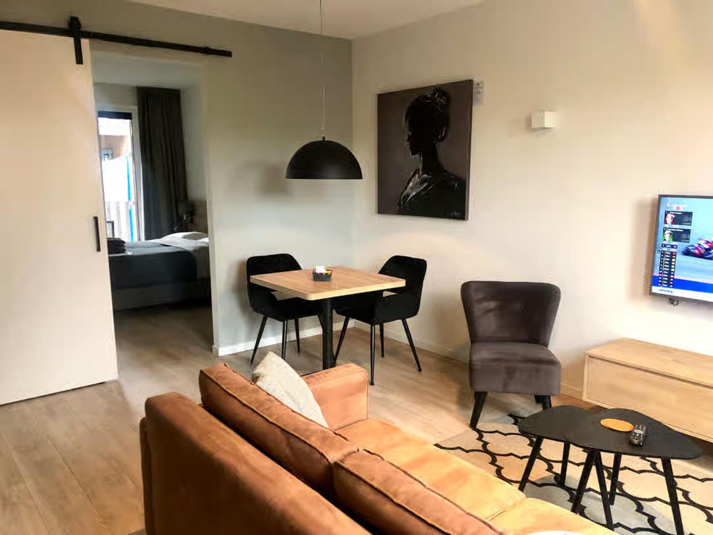 Un salon moderne avec un canapé, une table à manger et des chaises, un luminaire suspendu noir, un fauteuil gris, un téléviseur et un tableau au mur.