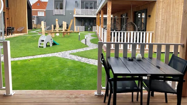 Eine Holzterrasse mit Tisch und Stühlen, ein Rasenplatz mit einer Rutsche und ein Gebäude mit einem Balkon.