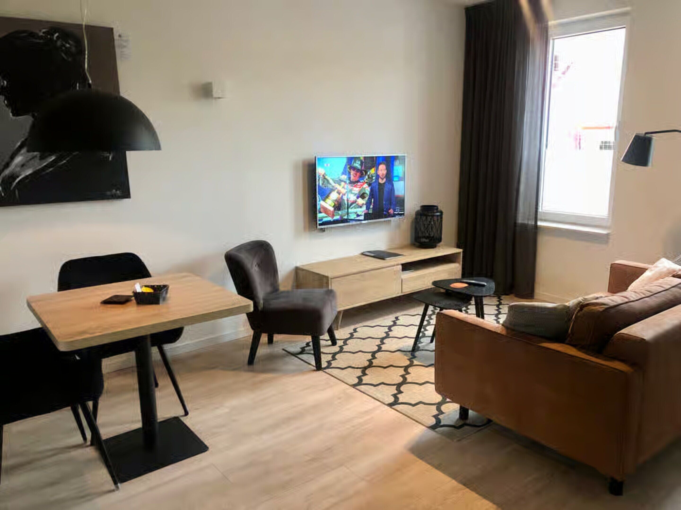 A modern living room with a wooden dining table, two black chairs, a brown sofa, and a TV on a wooden stand.