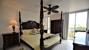 A four-poster bed with a canopy, a ceiling fan, a nightstand with a lamp, a dresser, and a view of the outdoors through a large window.
