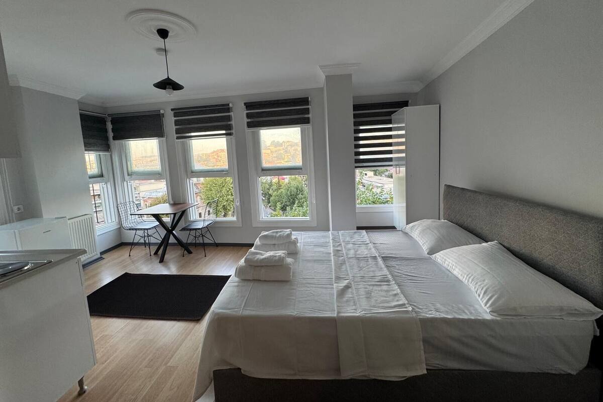 A modern bedroom with a large bed, a desk, and a chair. There are multiple windows providing a view of the outside.