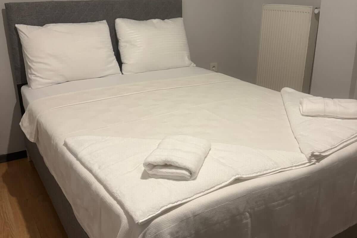 A neatly made bed with white linens and pillows, a grey headboard, and a radiator on the wall.