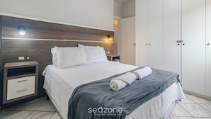 A neatly made bed with a gray blanket and a rolled towel, a bedside table with a lamp, and a wardrobe with a mirror. - Fully Furnished Apartments in Florianopolis - OPT (Florianópolis)