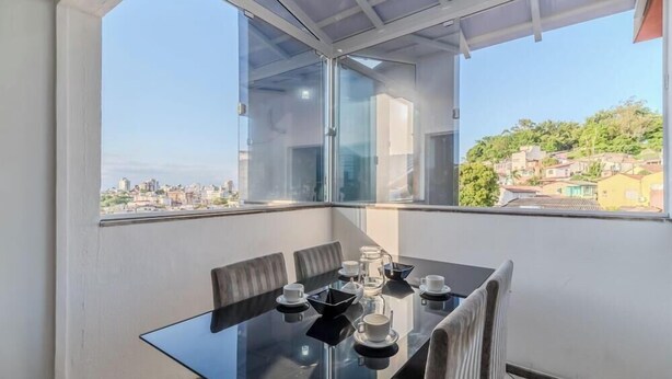 A modern dining area with a glass table and chairs, a coffee station, and a city view through large windows. - Fully Furnished Apartments in Florianopolis - OPT (Florianópolis)