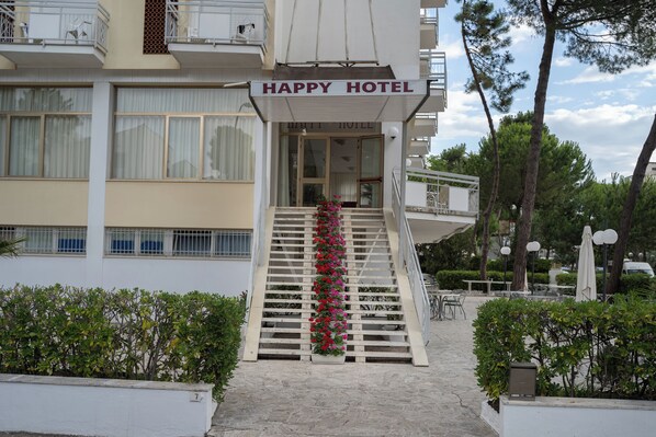 L’entrée d’un hôtel, avec une enseigne indiquant « HAPPY HOTEL », un escalier orné de fleurs rouges et des sièges installés à l’extérieur.