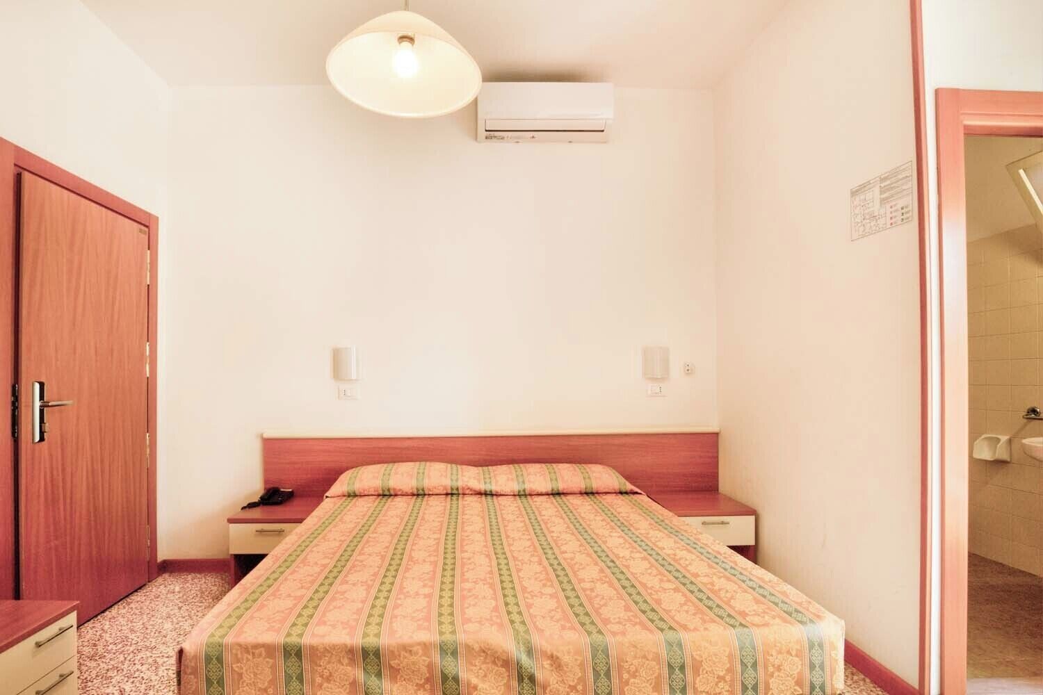 A small, single-bed room with a wooden headboard, a bedside table, an air conditioning unit, and a door to the left.