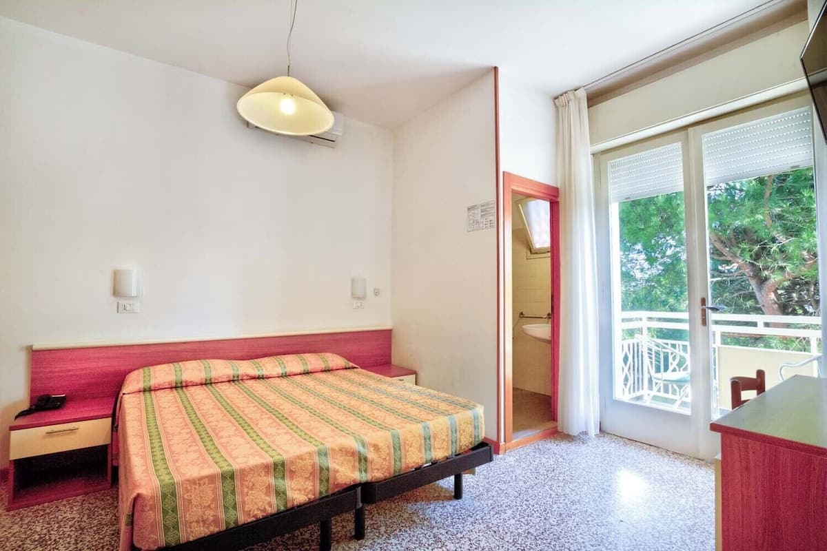 A bedroom with a bed, a nightstand, a desk, a window with blinds, and a balcony.
