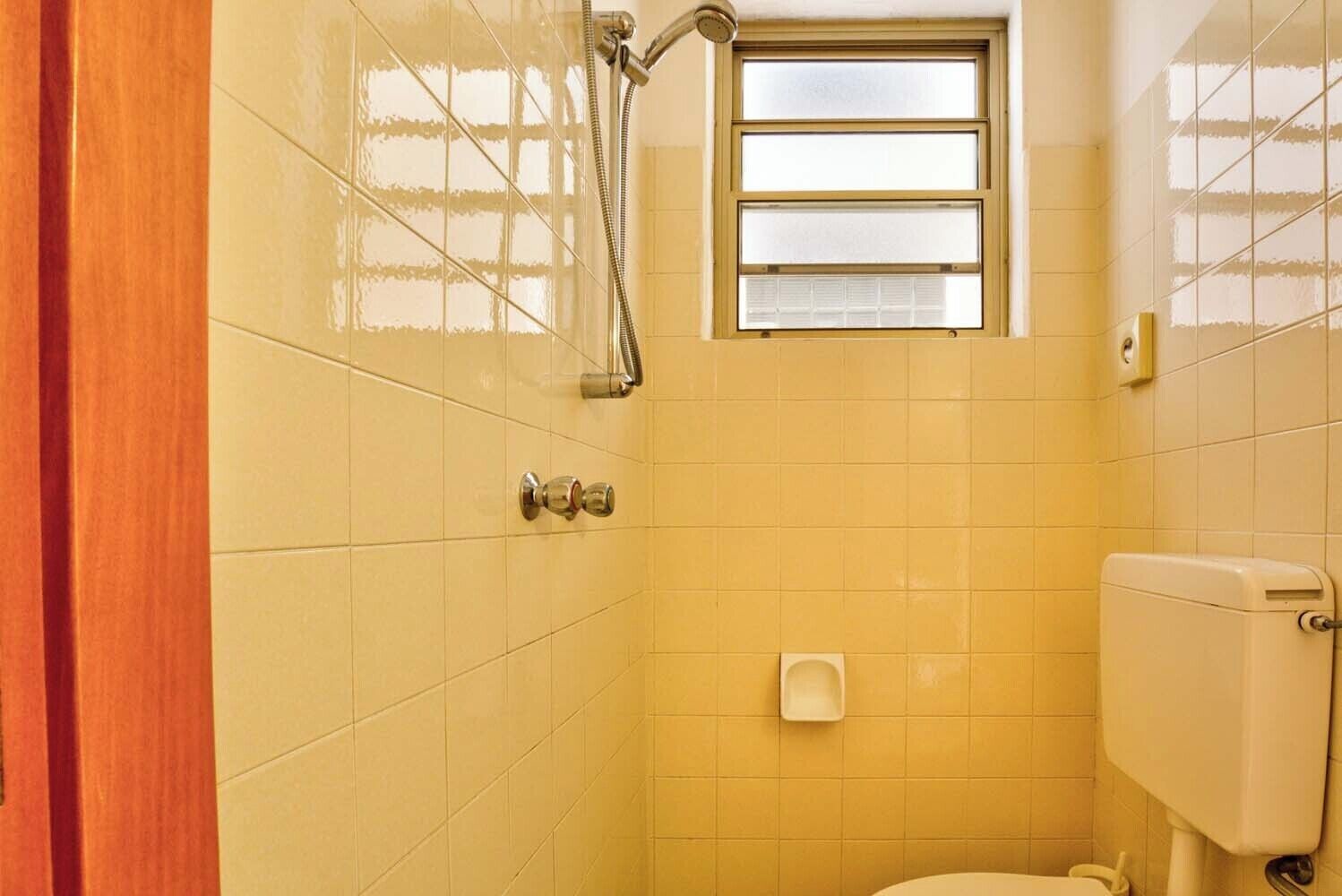 Un baño pequeño con baldosas amarillas, ducha, inodoro y ventana.