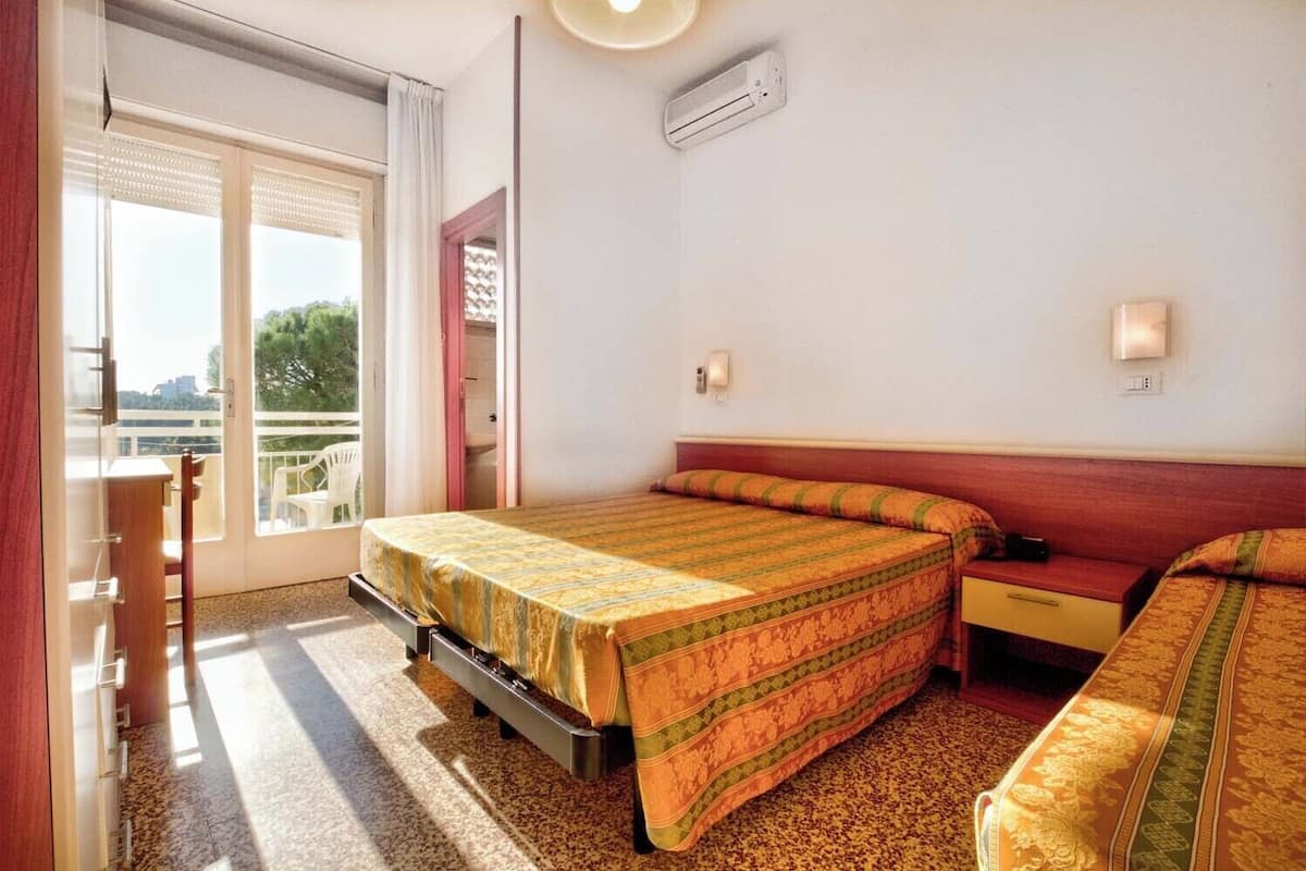 A hotel room with a bed, a desk, and a chair. There is a window with blinds, a door, and an air conditioning unit on the wall.
