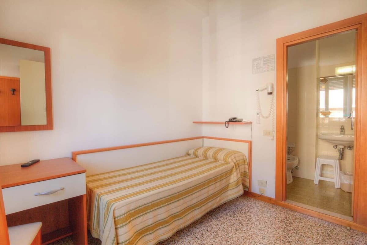 A small, single-bed room with a wooden desk, a mirror, and a bathroom visible through an open door.