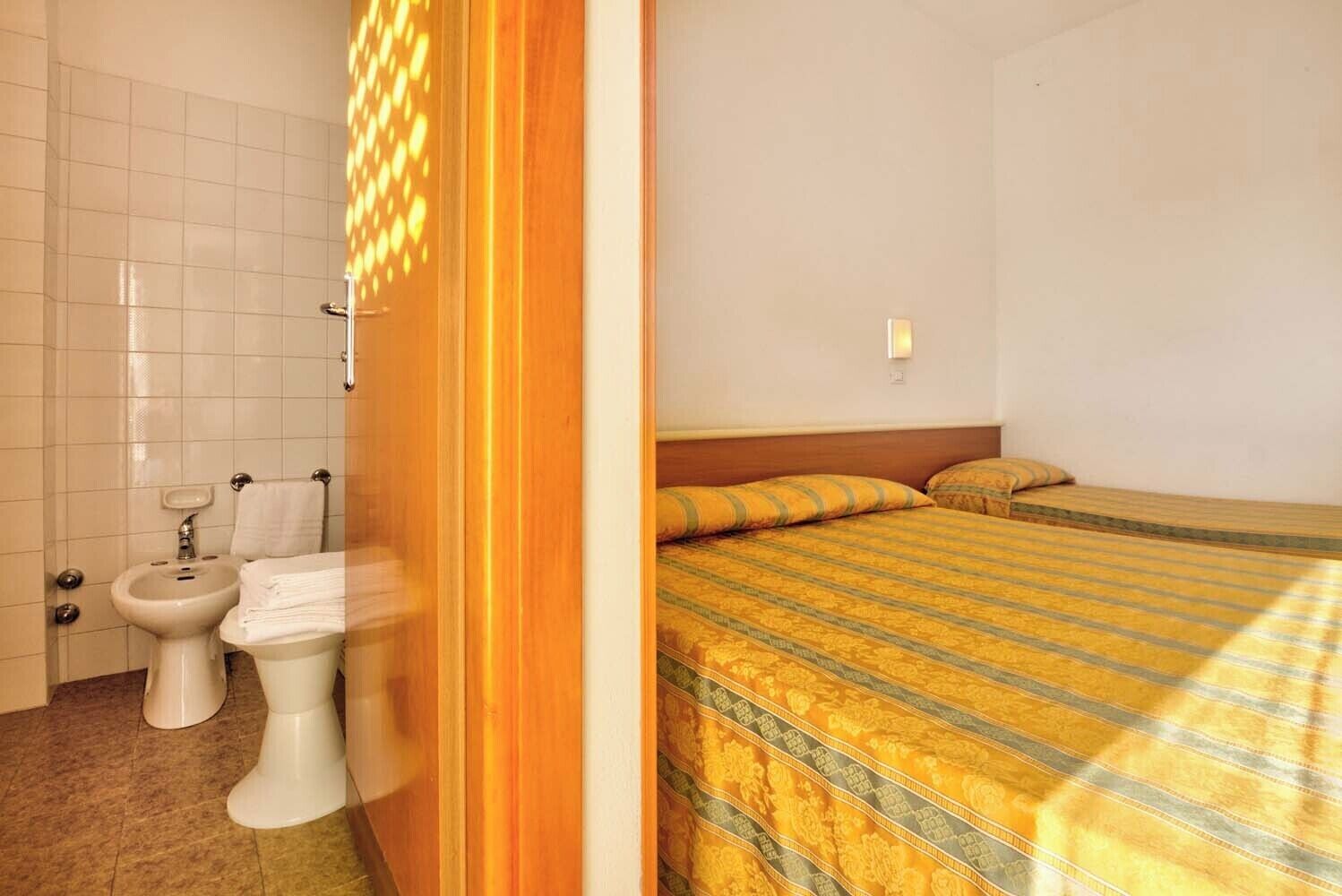 Ein Hotelzimmer mit Bett, Nachttisch, Fenster und einem Badezimmer mit Waschbecken und Toilette.