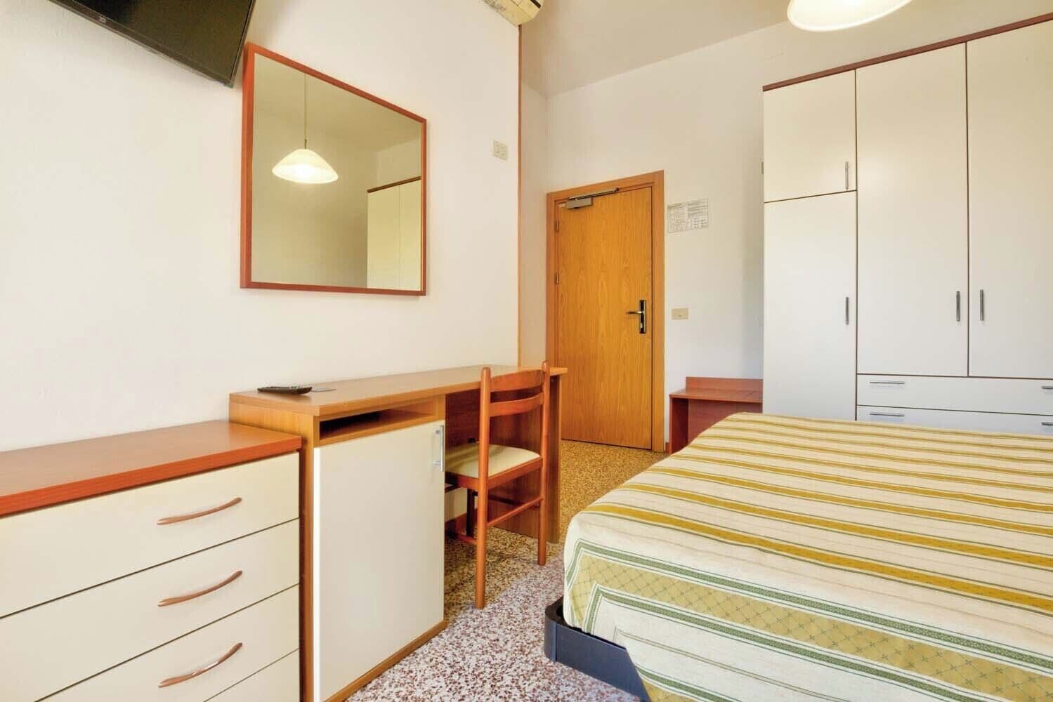 A bedroom with a bed, a desk, a chair, a wardrobe, and a mirror.