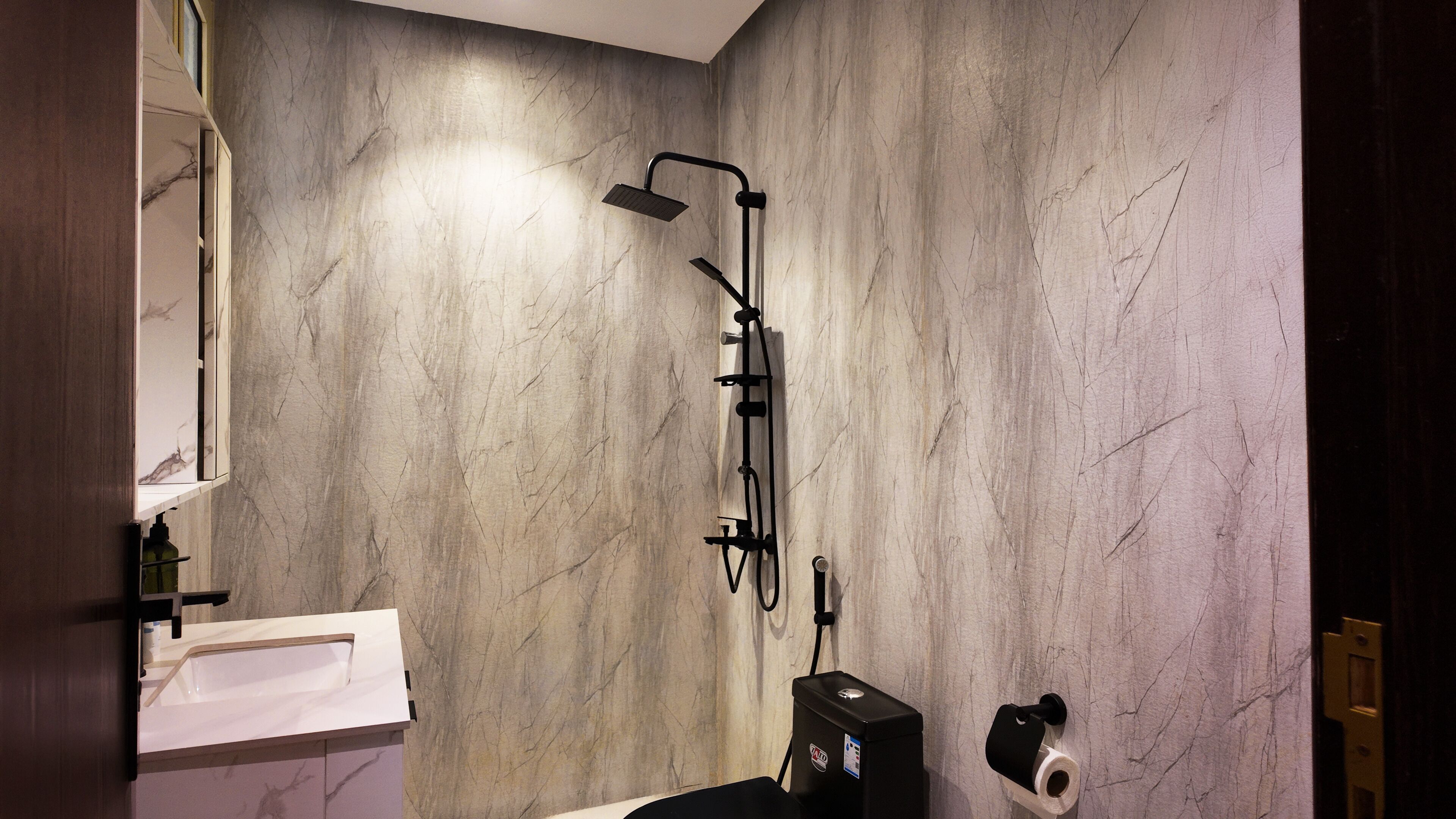A bathroom with a marble wall, a shower with a handheld showerhead, a toilet, and a paper towel dispenser.