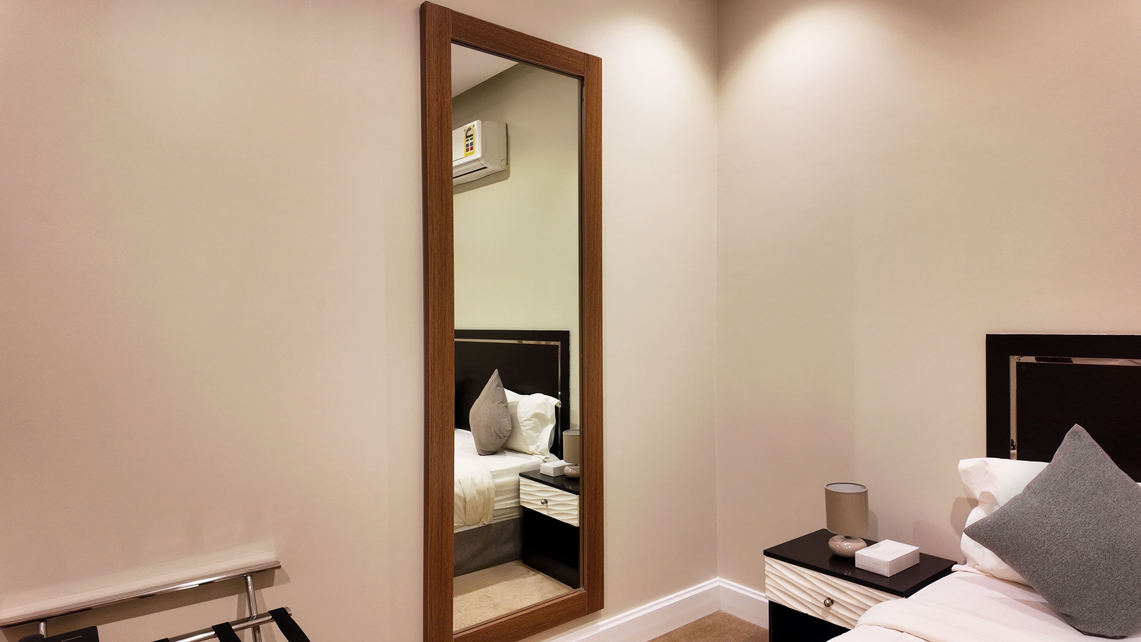 A hotel room with a large mirror, a bed with pillows, a bedside table with a lamp, and a nightstand with a lamp.