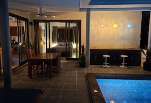 Ein moderner Poolbereich im Innenbereich mit Esstisch und Stühlen, einer Bar mit Hockern und einem Deckenventilator.