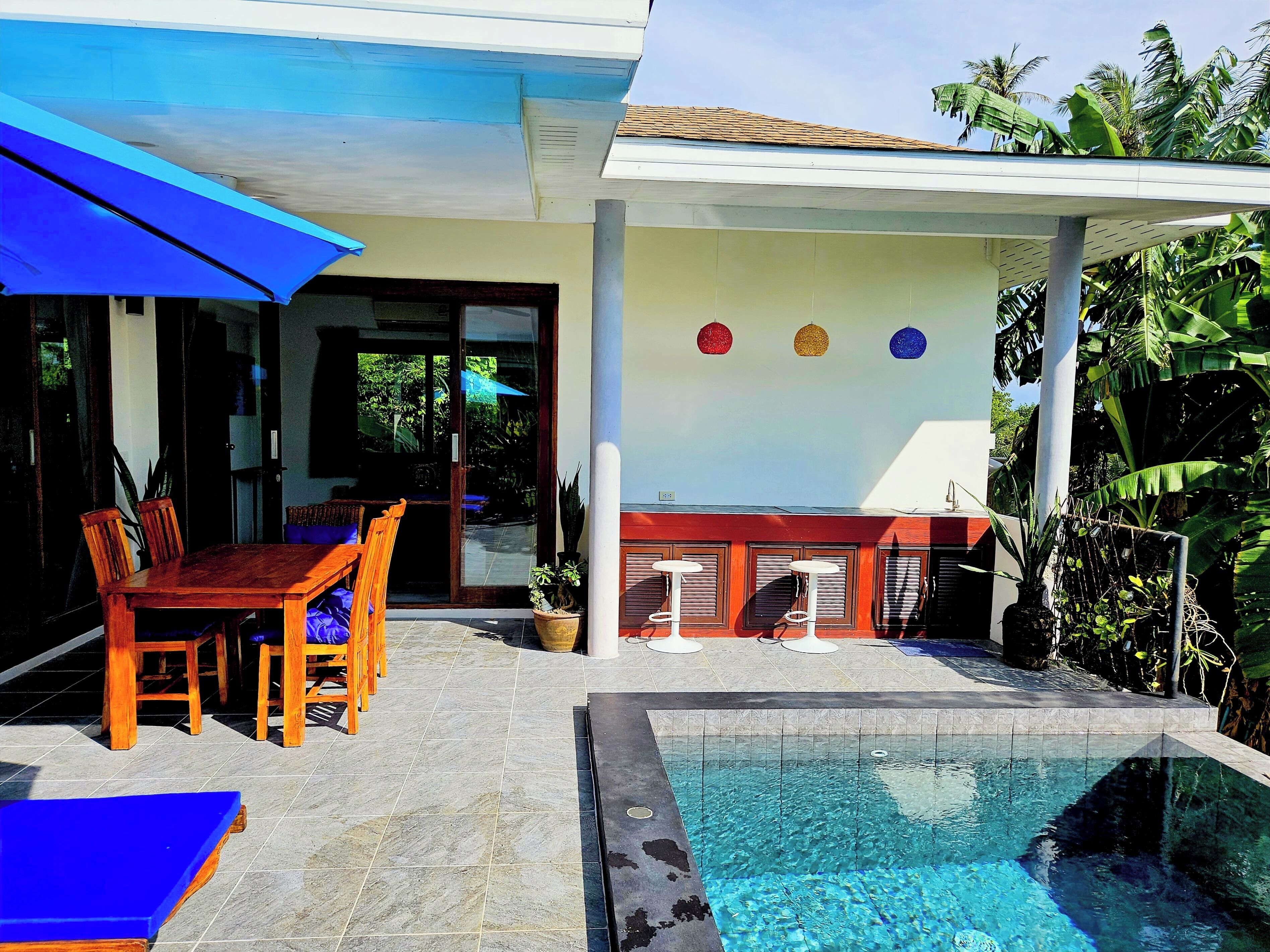 A pool area with a wooden table and chairs, a blue umbrella, and a tiled floor.
