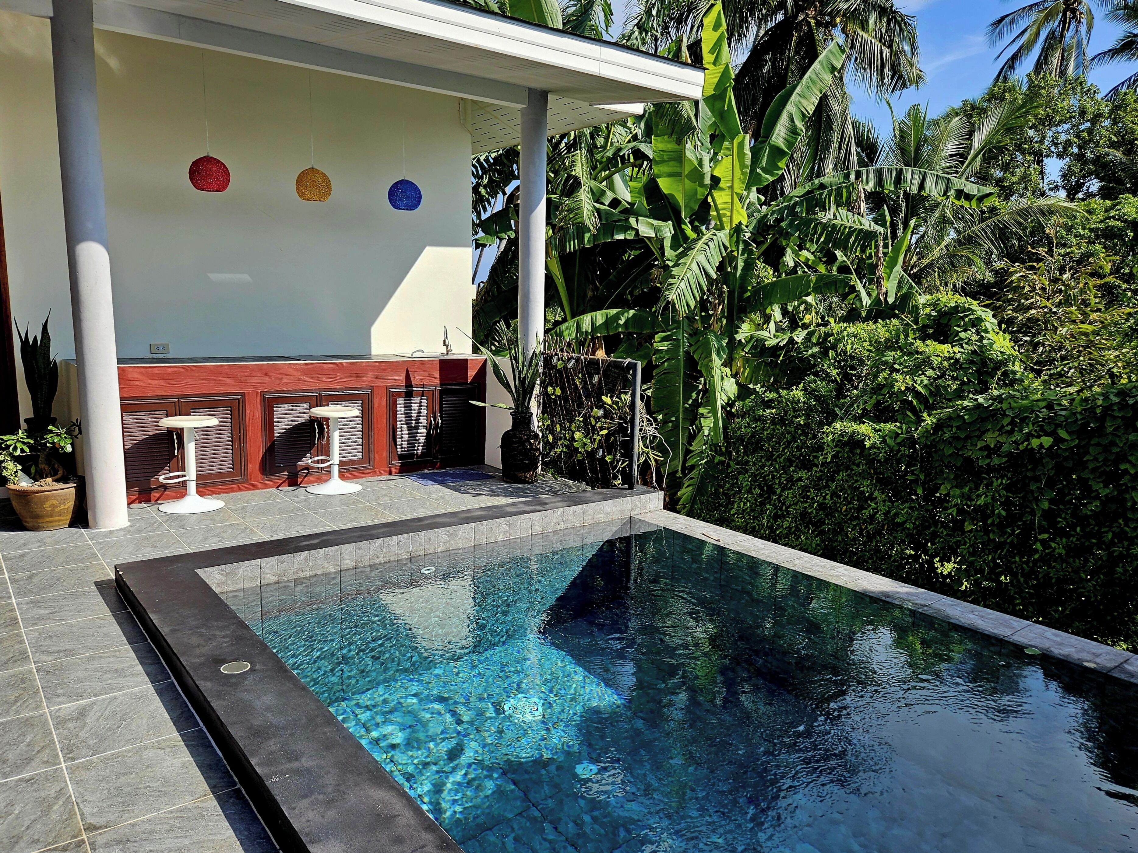 A swimming pool with a tiled deck, a covered seating area with a white chair, and lush tropical vegetation in the background.