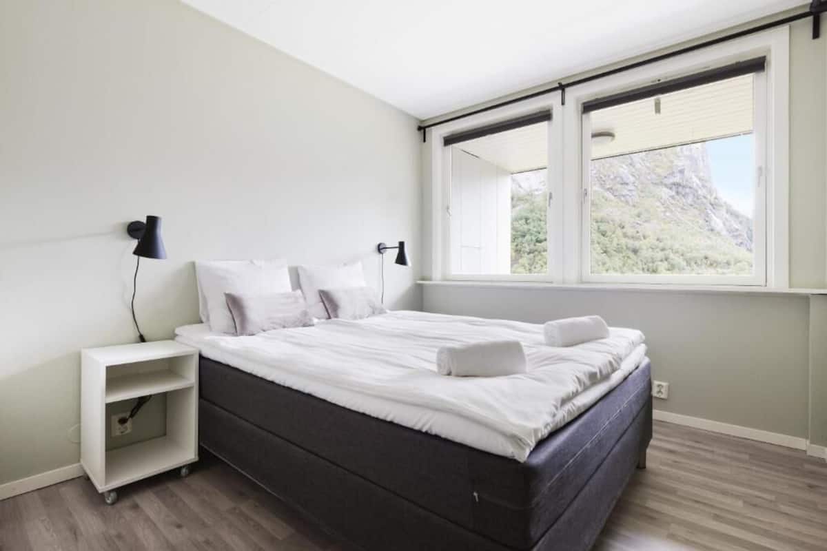 A modern bedroom with a bed, nightstands, and a large window with a mountain view.