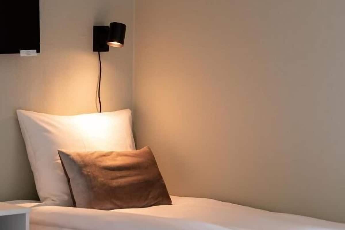 A hotel room with a single bed, a nightstand, a wall-mounted lamp, and a flat-screen TV.