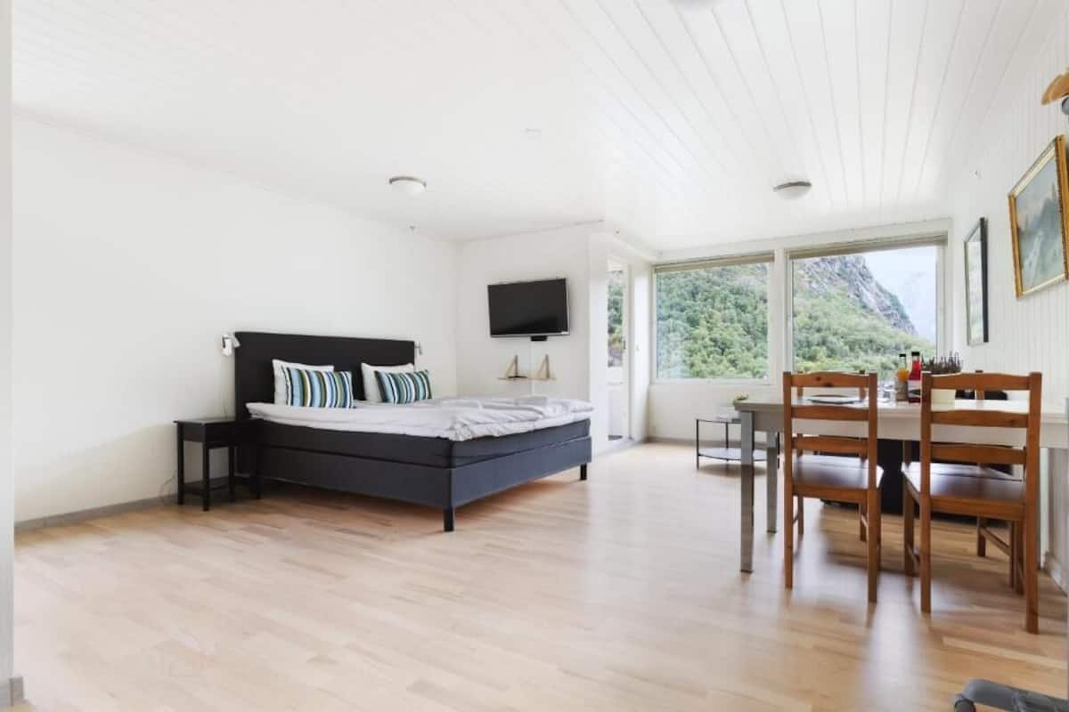 A bedroom with a bed, a TV, a dining table with chairs, and a view of greenery outside.