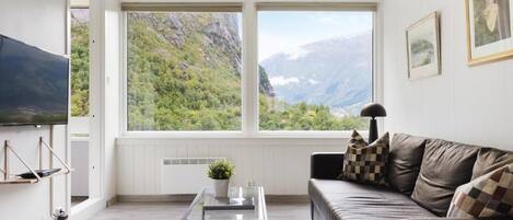 A modern living room with a large window offering a mountain view, a flat-screen TV mounted on the wall, a dark leather sofa, and a glass coffee table.