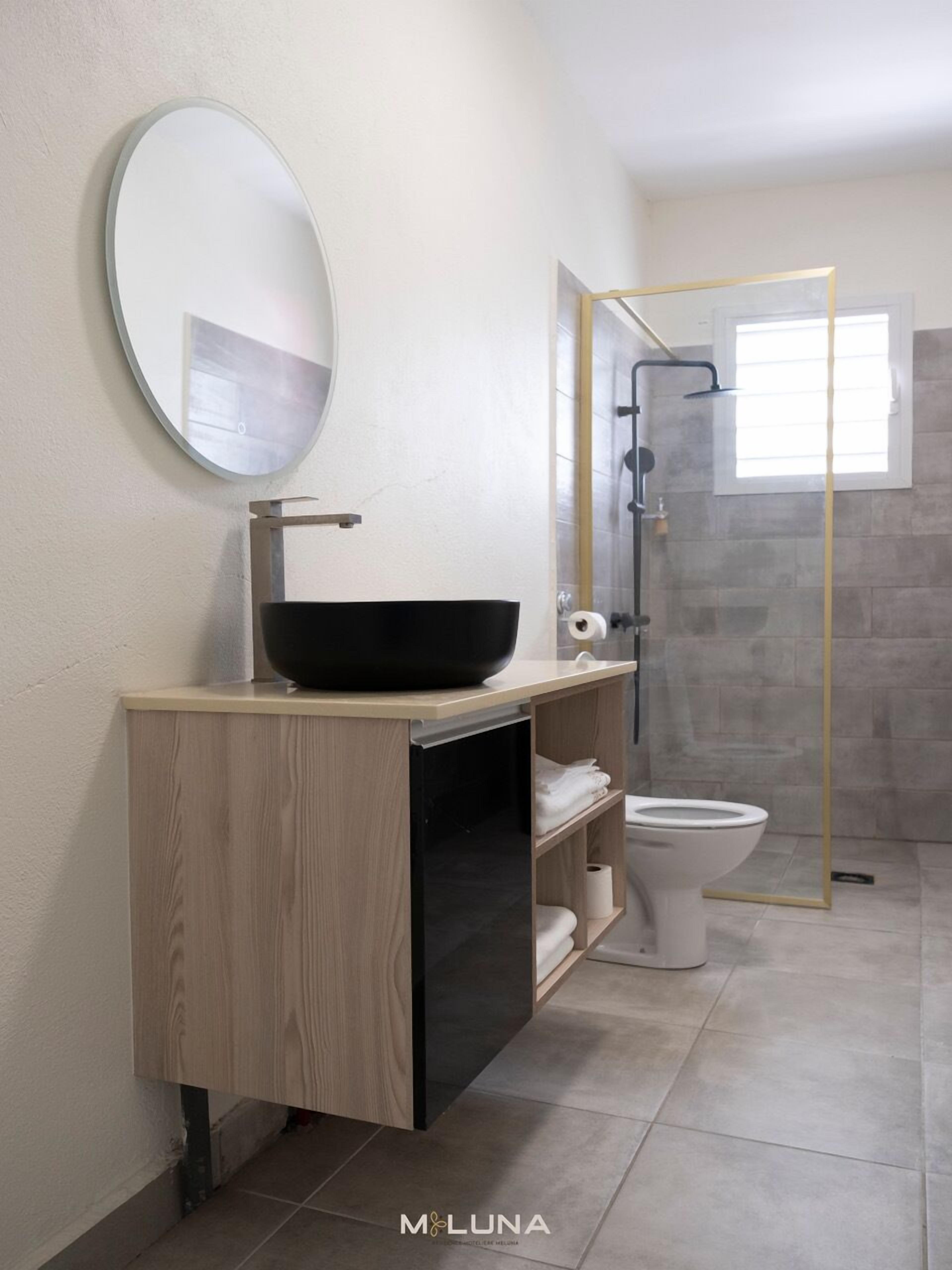 A modern bathroom with a wooden vanity, a round mirror, a black vessel sink, and a glass-enclosed shower.