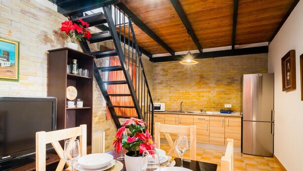 Un espace repas avec un mur en briques, un plafond en bois, un escalier, une cuisine équipée d’un réfrigérateur et un téléviseur.