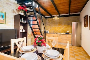Zona pranzo con parete in mattoni, soffitto in legno, una scala, una cucina con frigorifero e televisione.