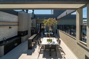 An outdoor patio with a dining table, chairs, and a flower arrangement. There is a grill and a seating area with a view of the sky. - APTO flat em condomínio com piscina B (Bombinhas)