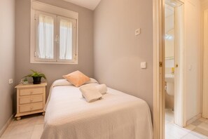 Een slaapkamer met een bed, een nachtkastje met een plant, een raam met gordijnen en een badkamer zichtbaar door een open deur.