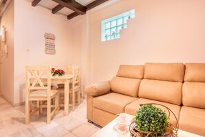A living room with a beige sofa, wooden dining table with chairs, and a small window with plants. - Apartment 'Lovely in Triana' with Wi-Fi and Air Conditioning (Sevilla)
