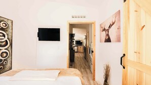 Ein Schlafzimmer mit einem Bett, einer Holztür, einer durchgehenden Tür erkennbaren Küche und einem an der Wand befestigten Fernseher.