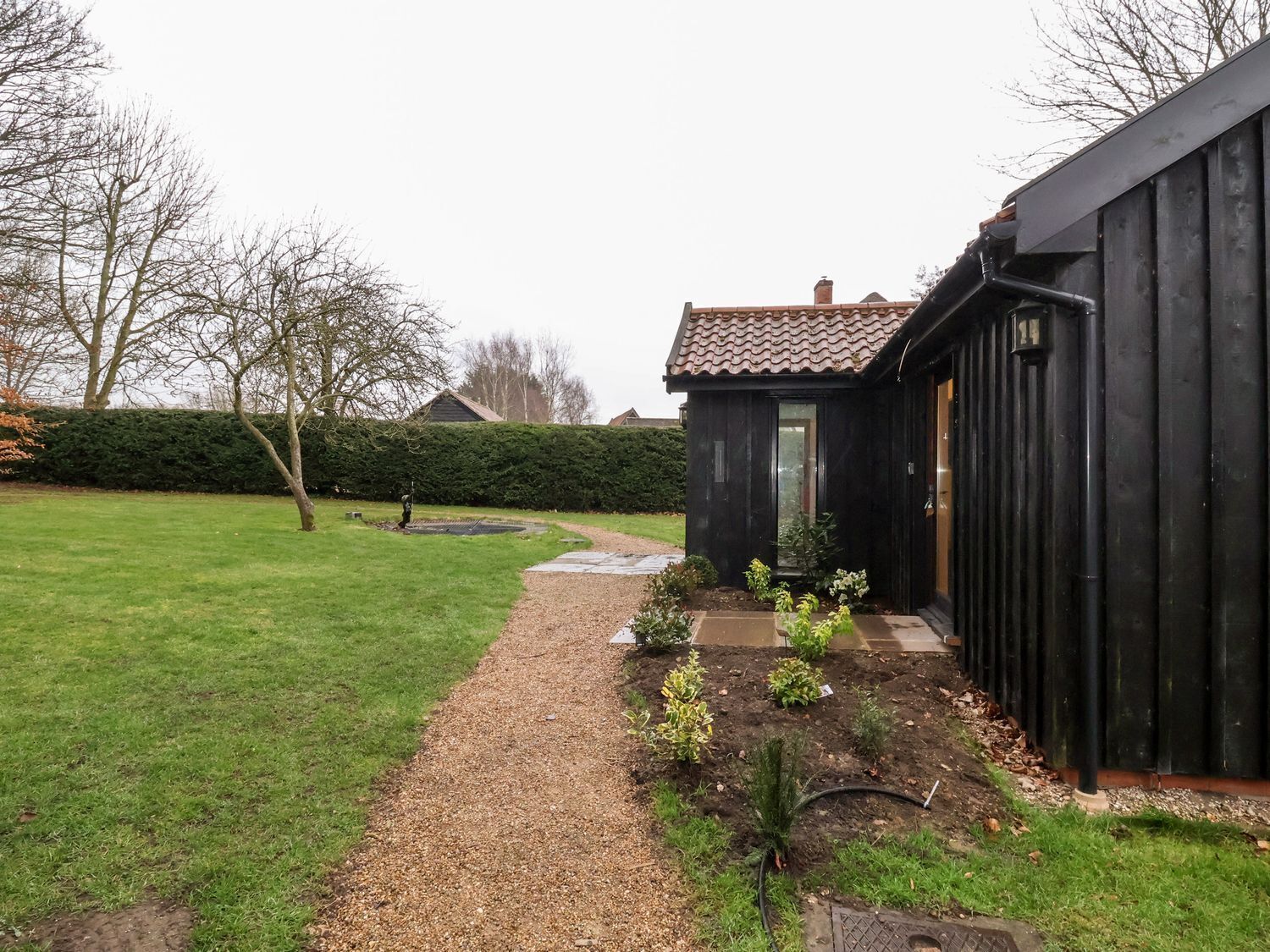 A pathway leading to a black building with a tiled roof, surrounded by a well-maintained lawn and a hedge.