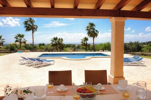 Zona pranzo a bordo piscina con vista su piscina, palme e paesaggio in lontananza.
