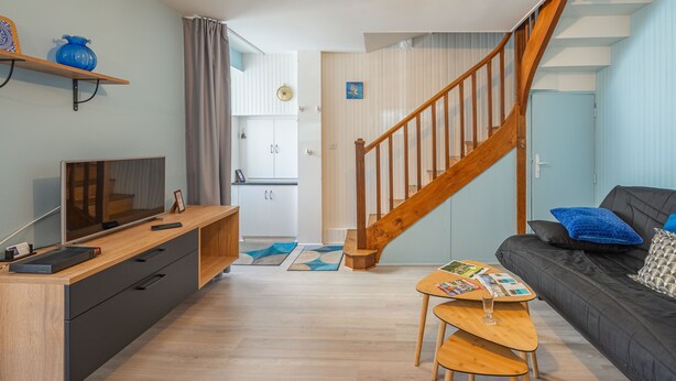 A modern living room with a wooden staircase, a flat-screen TV, a sofa with cushions, a coffee table, and a small dining area. - Furnished Apartment in Townhouse, City Center Location (Jonzac)