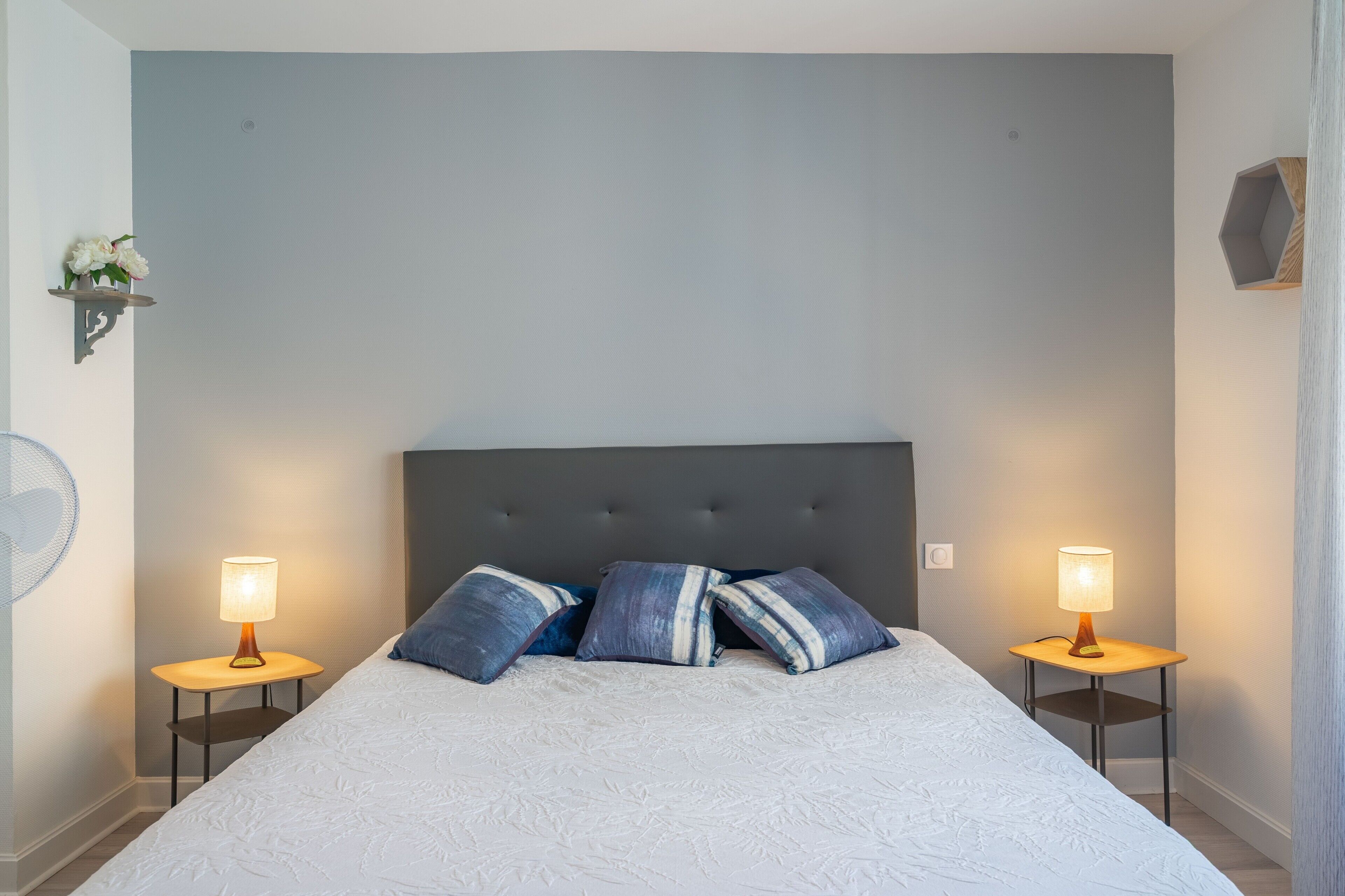 Une chambre à coucher avec un grand lit, deux tables de chevet avec des lampes, une tête de lit sombre et une étagère murale décorée de fleurs.
