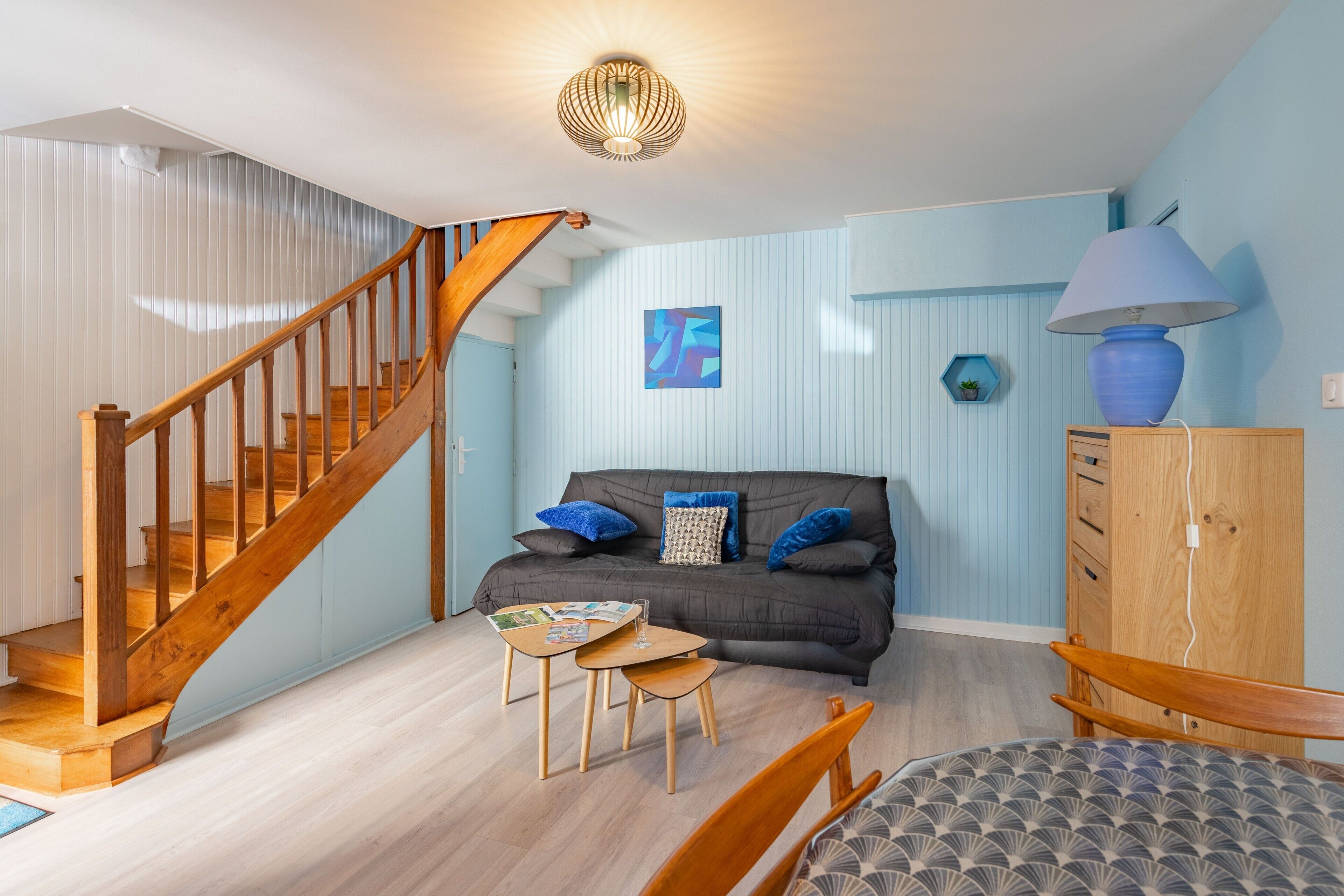 A room with a wooden staircase, a dark-colored sofa with blue cushions, a small wooden table, and a lamp on a wooden cabinet.