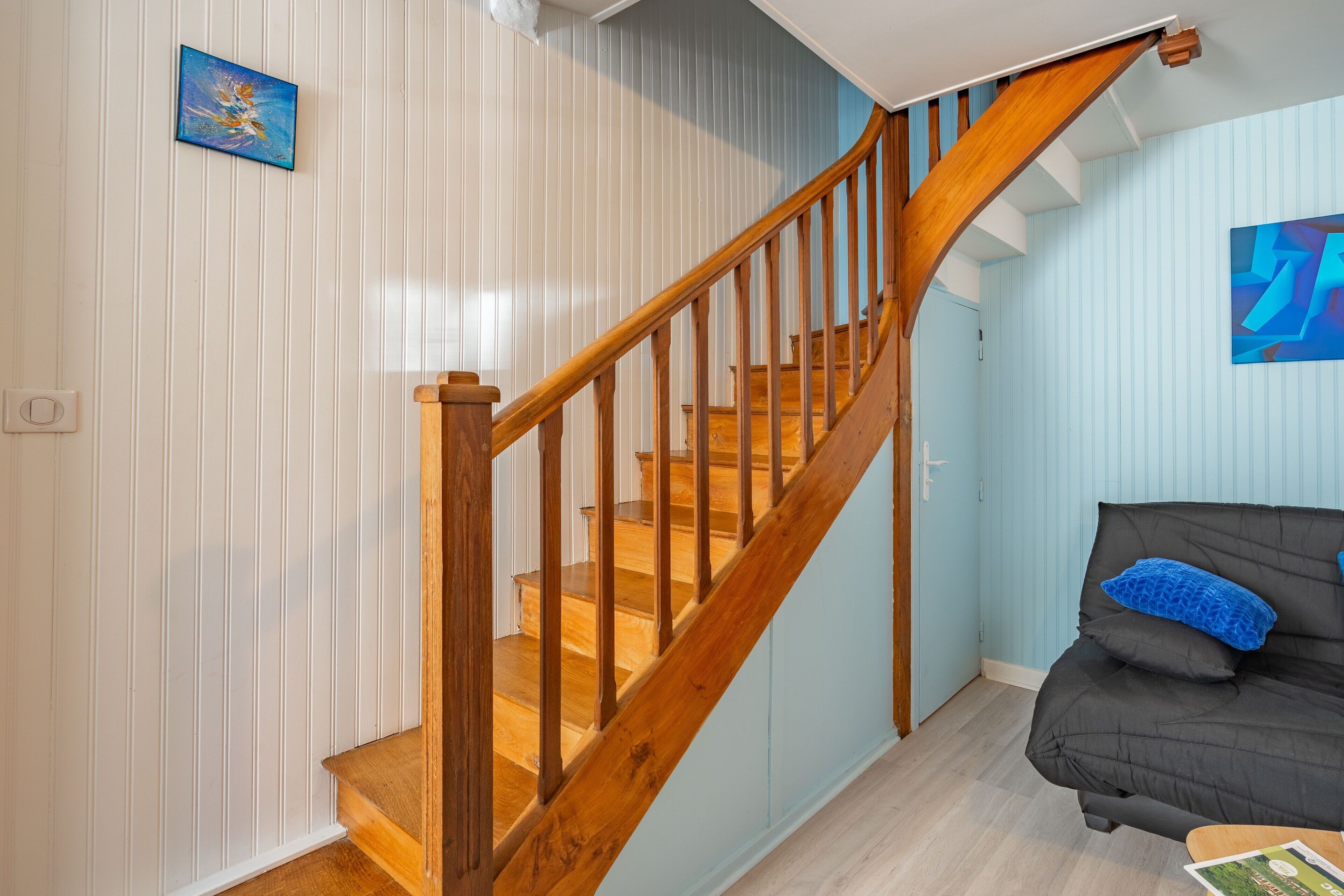 Een trap met houten leuningen die leidt naar een plateau, een donkergroene bank met een blauw kussen, en een lijst met een afbeelding aan de muur.