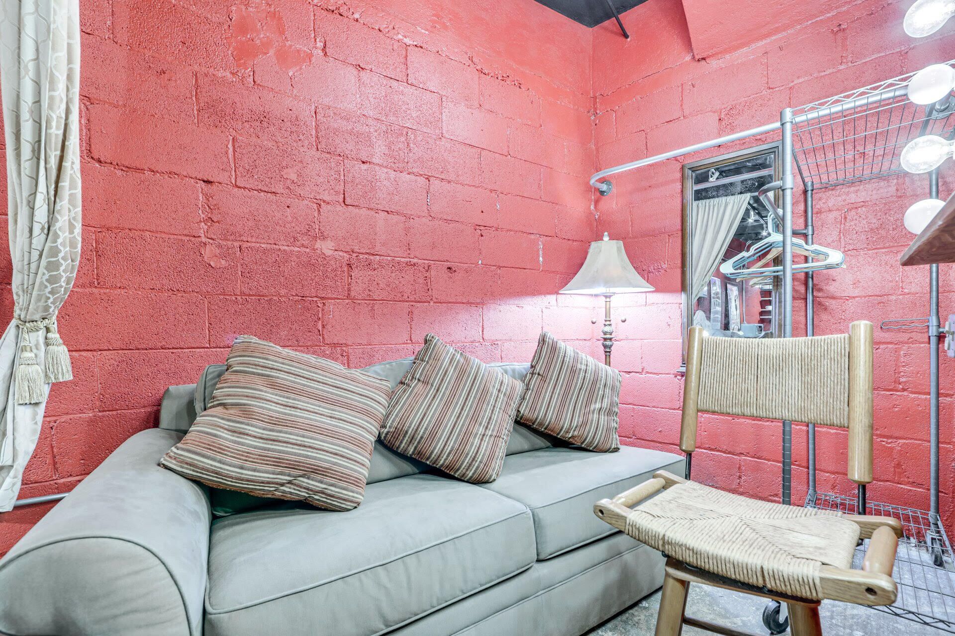 Una habitación con pared de ladrillo rojo, un sofá gris con almohadones a rayas, una silla de madera, un perchero metálico y una lámpara.