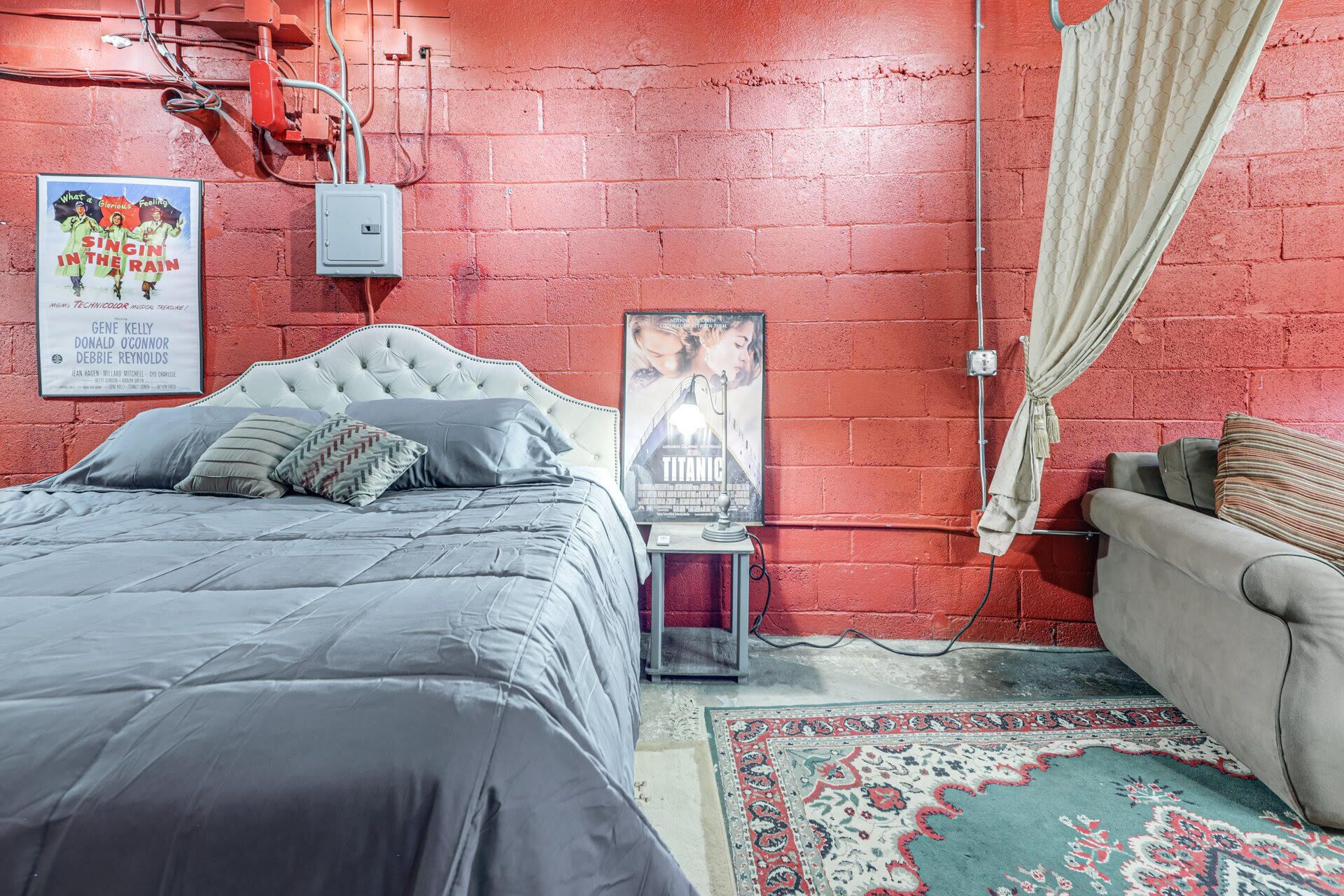 A bedroom with a red brick wall, a bed with grey bedding, a poster of 'Titanic', and a small bedside table with a lamp.