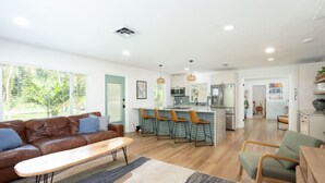 Un salon moderne avec un canapé, une table basse et des fauteuils, une cuisine avec des tabourets de bar et un coin repas.