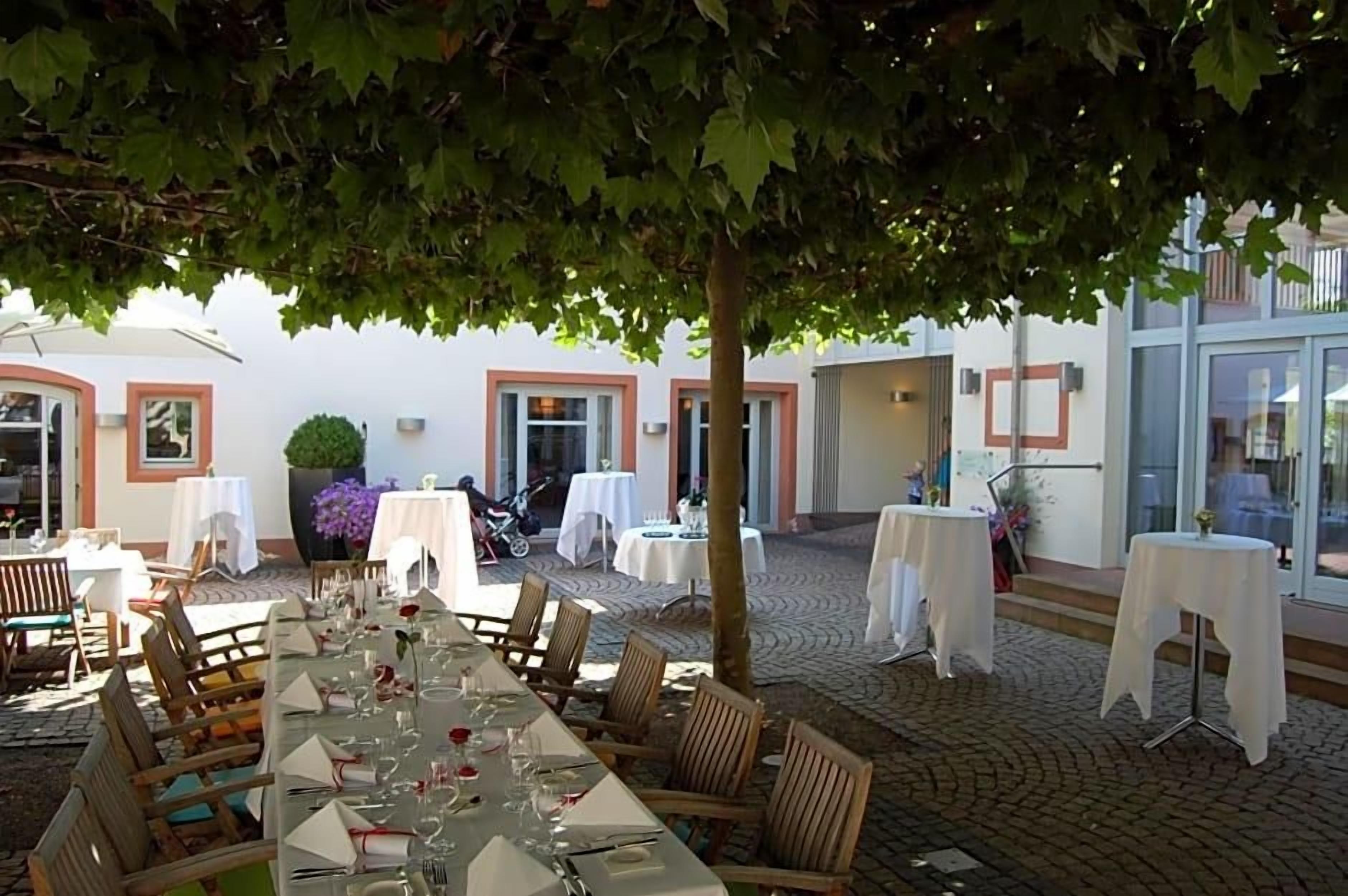 Un espace de restauration en plein air, avec des tables dressées pour un repas, entouré d’un bâtiment dont le sol est recouvert de carreaux et sur lequel se trouve un arbre.