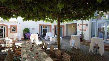 Un espace de restauration en plein air, avec des tables dressées pour un repas, entouré d’un bâtiment dont le sol est recouvert de carreaux et sur lequel se trouve un arbre.