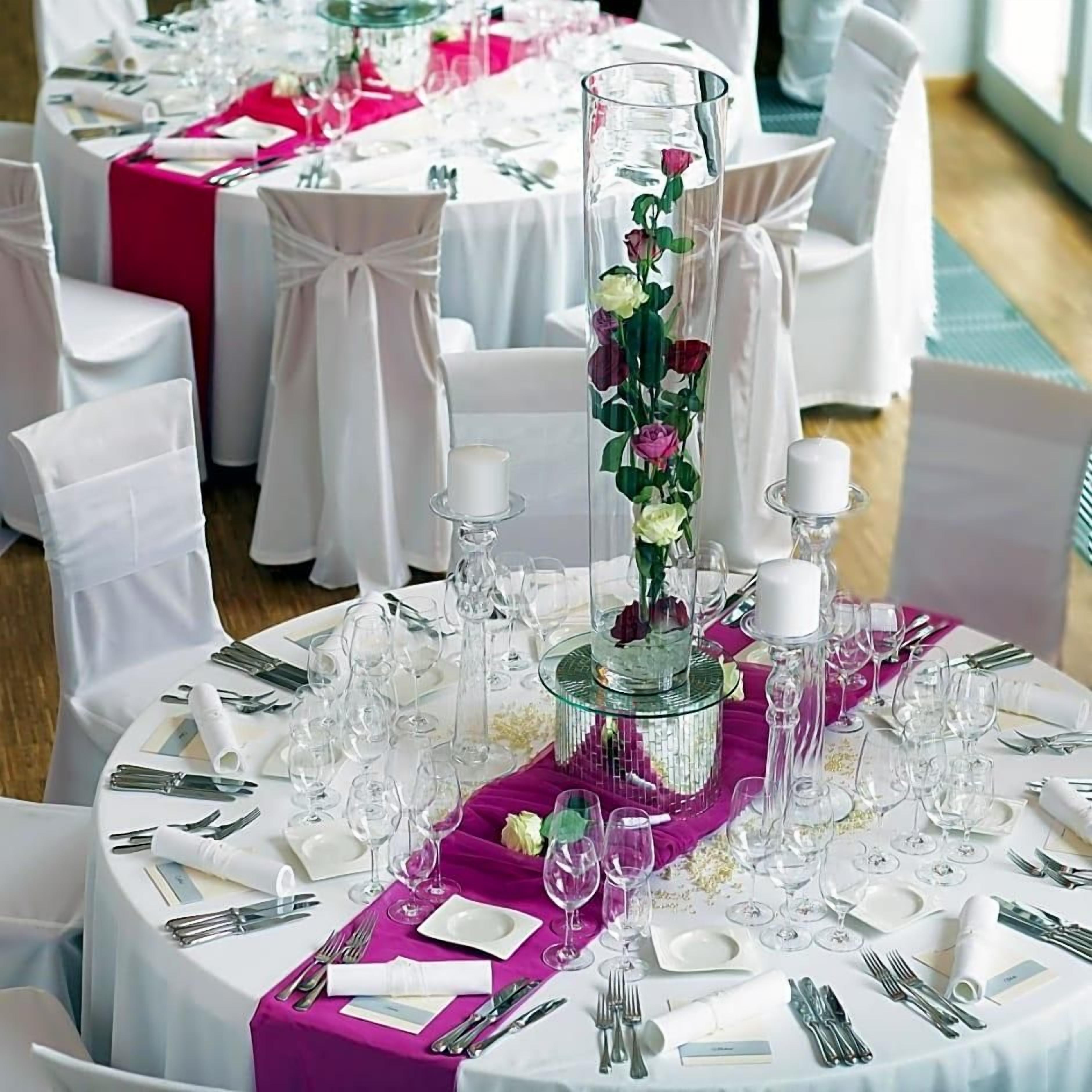 Una mesa redonda de comedor con manteles blancos, centros de mesa rosados y sillas blancas.